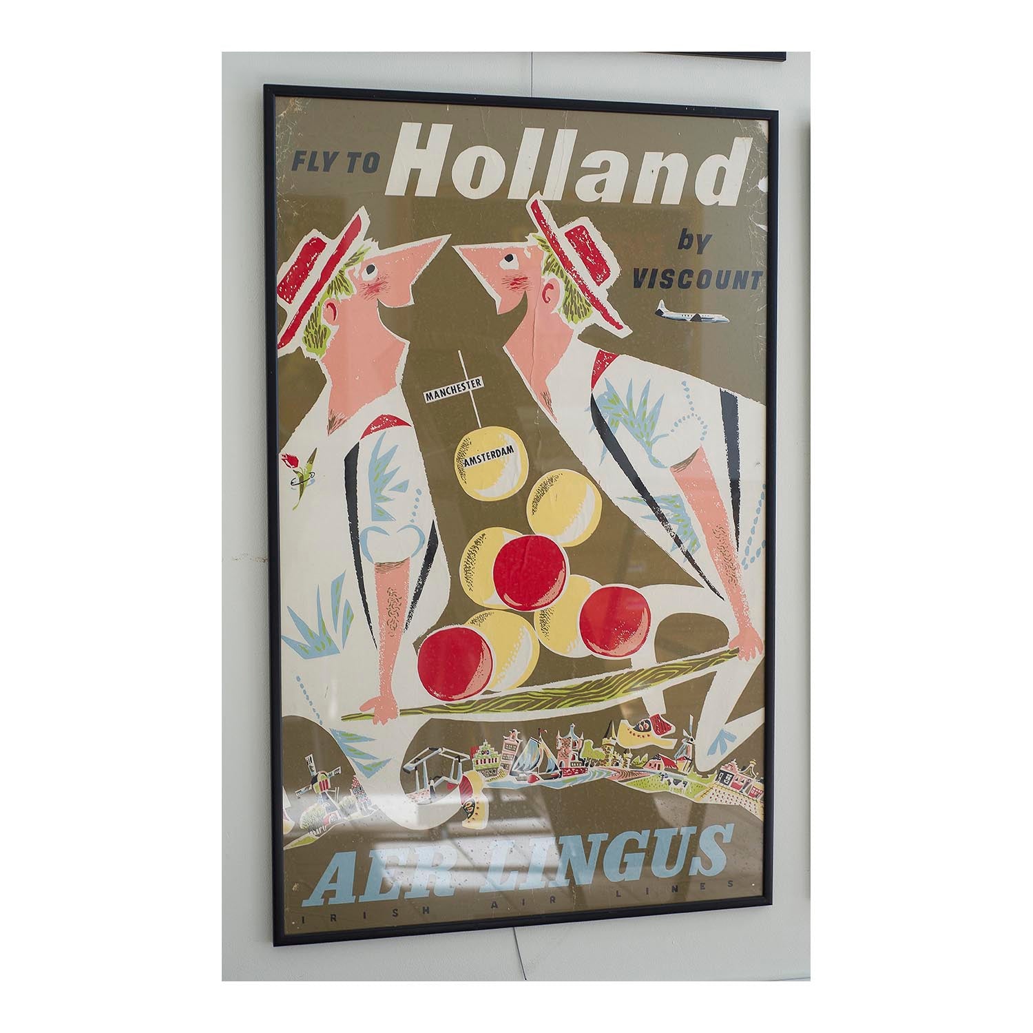 An original travel poster, Holland by Viscount, published by Aer Lingus, c. 1957-58. The poster publicises flights from Manchester to Amsterdam with a depiction of which is included flying over two Dutchmen, precariously delivery a batch of circular Edam cheeses.