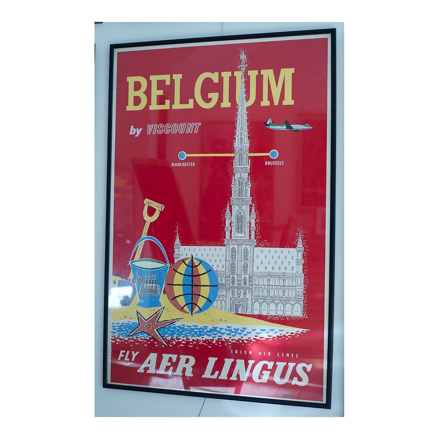 An original travel poster, Belgium by Viscount, published by Aer Lingus, c. 1957-58. The poster publicises flights from Manchester to Brussels, with a plane flying over Grand Place, Brussels, with beach toys and sand in the forefront.